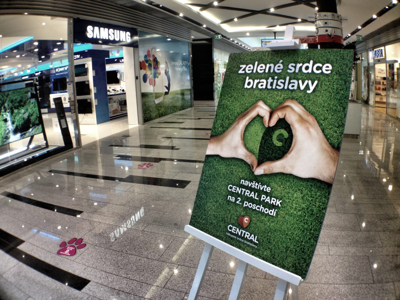 Plagát „Zelené srdce Bratislavy" na stojane v pasáži centra: ruky zložené do tvaru srdca na trávniku, pod nimi výzva navštíviť Central Park na 2. poschodí a logo Central s podtitulom nákupné srdce Bratislavy